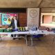 A student standing behind two white tables filled with surplus food and next to a banner with the Food Waste Cafe logo under the arches of Falmer House