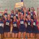 The Netball team in their uniforms celebrating their victory holding the cup and a yellow sign identifying them as varsity series winners