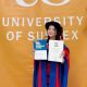 Dr Bendangyangerla Imchen in her doctoral robes holding up her degree certificate and Spirit of Sussex Award