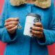 A woman holding the returnable cup in one hand and a token in the other, she's in a blue coat with a red background