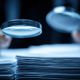 out of focus hands holding two magnifying glasses above a pile of papers