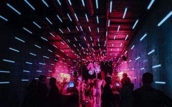 Young people dancing in a narrow and long club. Thin long lamps on the walls and ceiling guide the observer to the end of the room where the dancefloor is illuminated by a pink and purple light