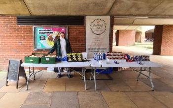 A student standing behind two white tables filled with surplus food and next to a banner with the Food Waste Cafe logo under the arches of Falmer House