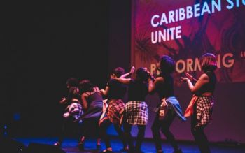 Students dancing in a line on the stage of the Attenborough Centre for the Creative Arts. Behind, a partial text reads Caribbean Stu Unite