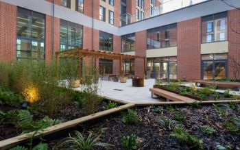 View of the Student Centre courtyard from behind two raised plant beds in an L shape. The garden includes benches near raised plant beds and a covered area on the other side of the patio