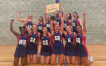 The Netball team in their uniforms celebrating their victory holding the cup and a yellow sign identifying them as varsity series winners