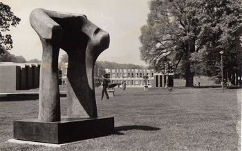 Black and white picture of a stone sculpture standing on a pedestal on campus.