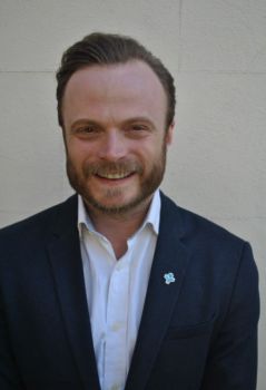 Bearded man smiling at the camera in a white shirt and a blue jacket with a light blue pin on his lapel representing a person with open arms