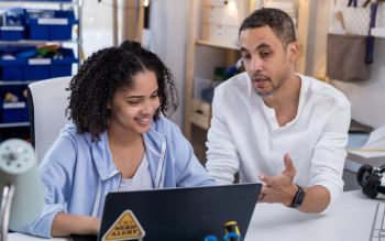 young female student writing on her laptop while a man is explaining something to her