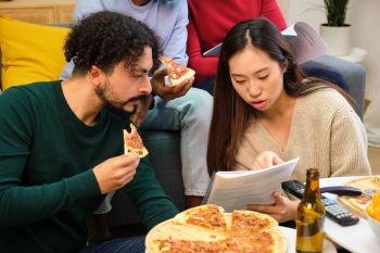 Two young people read a document whilst eating pizza.