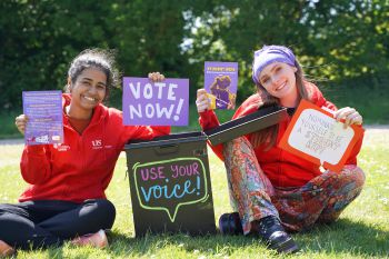 Two women wearing red Students' Union hoodies hold up signs and leaflets encouraging people to get involved.