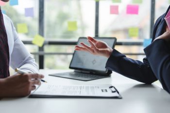 A hand makes notes on a clipboard whilst across a desk another hand gestures explaining something.