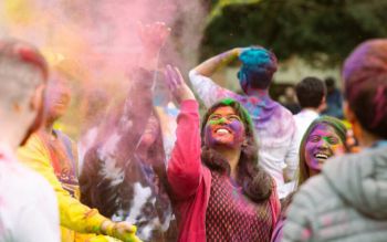 Group of students joyfully celebrating Holi, their faces and clothes covered in vibrant colors. They are laughing and throwing coloured powder into the air, creating a cloud of hues.