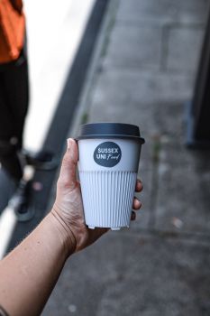 A caucasian woman's hand holding a returnable cup with a label that says 'Sussex Uni Food'