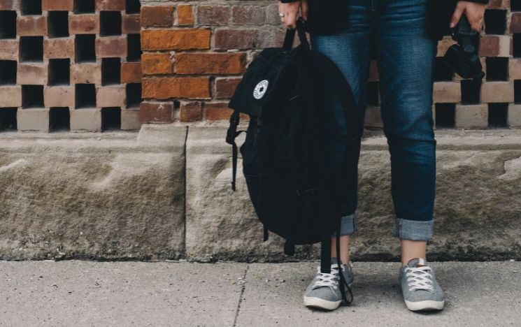 Legs of a person standing by a wall carrying a backpack in their hand