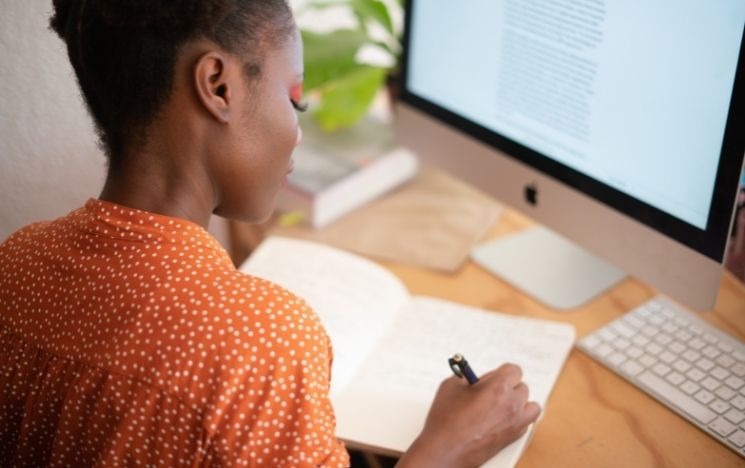 Black woman writing on her notebook in front of a computer