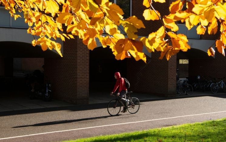 Cyclist in a red raincoat riding his bike past Pevensey 1 building and a tree with yellow leaves