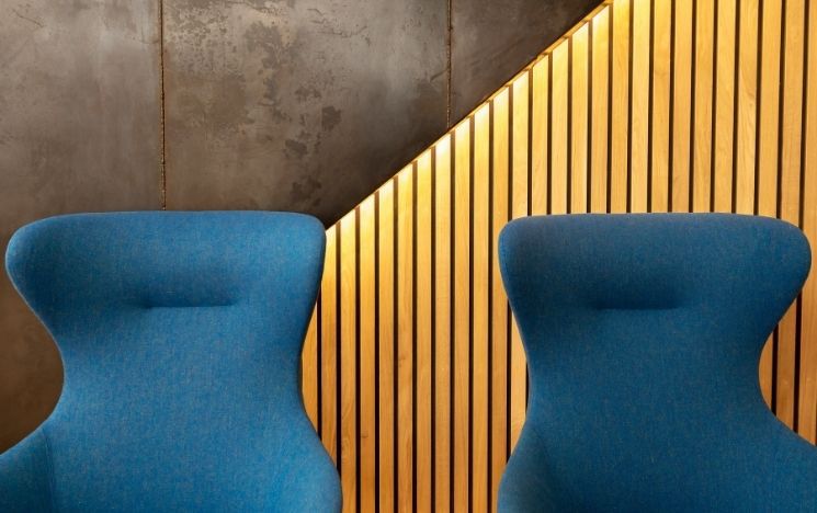 Two blue modern-looking armchairs. The wall behind is divided into two section diagonally; the one on the left look like an industrial iron structure and the one of the left is covered with thin vertical wooden panels