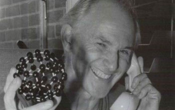 Black and white picture of Harry Kroto smiling while he speaks on the phone with the Nobel Prize panel. He's holding a figure of the C60 molecule