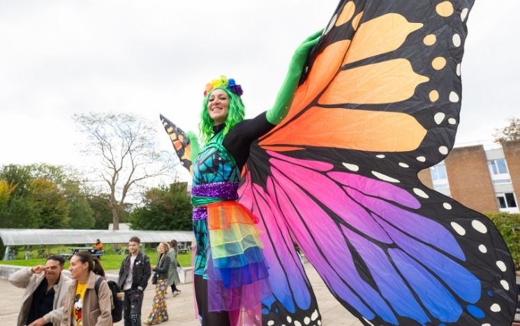 A woman stands on stilts in a colourful outfit with huge, multicoloured butterfly wings and a green wig smiling as students walk past her.