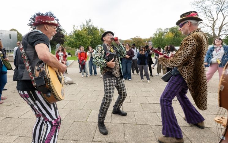  Three musicians with guitars and a harmonica perform to a crowd of students near the business school.
