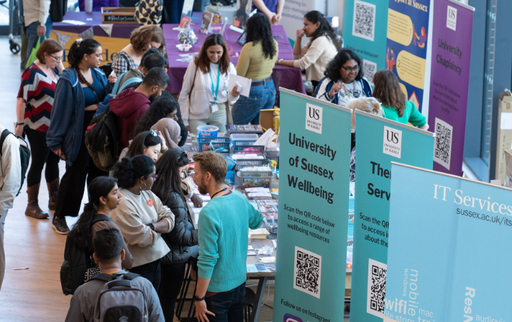 Busy Welcome Week event with students and staff at informational booths, featuring banners for University of Sussex Wellbeing and IT Services.