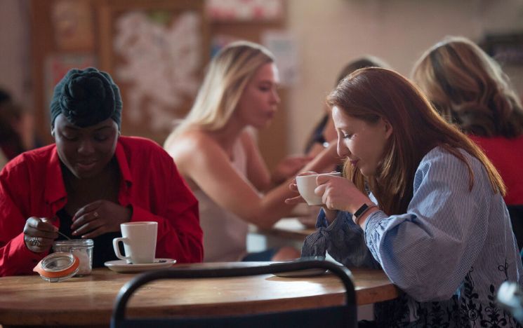 Two students at a coffee shop on campus. One of the is blowing on her hot coffee while her friend is adding sugar to her cup