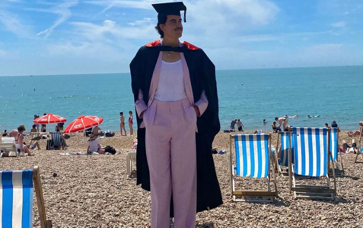 Pablo in graduation clothing on Brighton Beach with deck chairs and people in the background