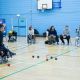 In the Sports Centre, a student on a chair is throwing a small blue ball at other blue and red balls on the floor in front of he. Next to her, four students on wheelchairs or chairs wait their turn. Opposite a man on wheelchair encourages her to play