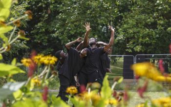 Four men in black t-shirts dancing on a lawn with trees and foliage in the background and foreground.